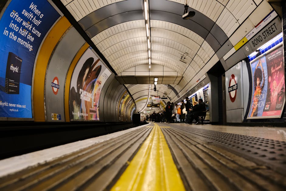 A low-angle view inside an underground London tube station, specifically at the platform in front of the Tunnel entrance with clear yellow safety lines running along the edge. The curved ceiling is li