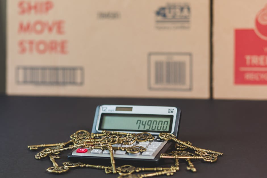 A flat-lay image features various items related to house removals and packing sitting on a light brown surface. Visible objects include a black marker, a roll of black tape, a silver key attached to a keychain with a house-shaped charm, and a calculator. Part of a white tablet or document with coloured graphics is seen on the right side. A portion of a yellow tape measure and red measuring tape are also present. The cardboard surface displays red printed instructions reading 'FOLD TO HOLD' and icons illustrating packing and moving processes, such as boxes and circular arrows. These objects and markings collectively suggest a preparation or planning stage for home relocation, showcasing items involved in packing, securing, and transporting household goods. The scene emphasizes the organized and professional approach of Man With a Van Holloway during furniture transport and removal services.