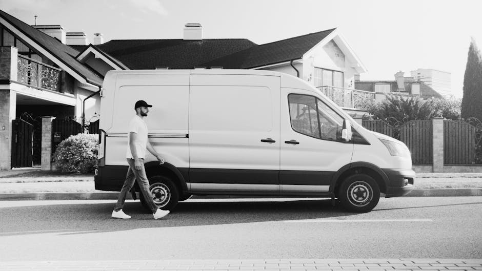 A man wearing a light grey cap, black gloves, a blue long-sleeved shirt, and a black padded vest stands beside an open van in a residential driveway during daytime. The van has a wooden panel inside,