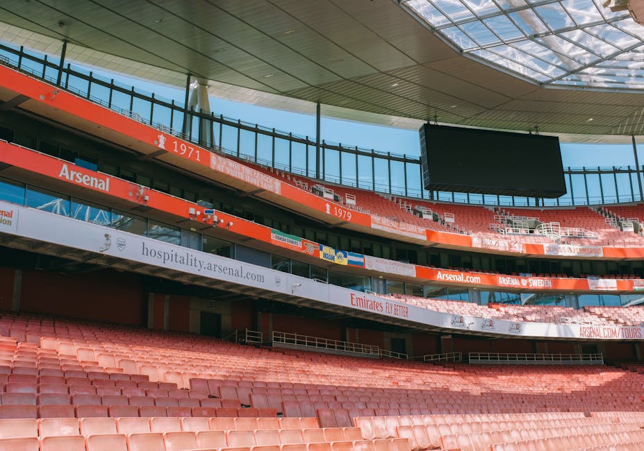 A wide-angle view of Emirates Stadium during sunset, showing its modern, curved glass facade and large digital display screen featuring an Arsenal football team graphic with players in red kits and th