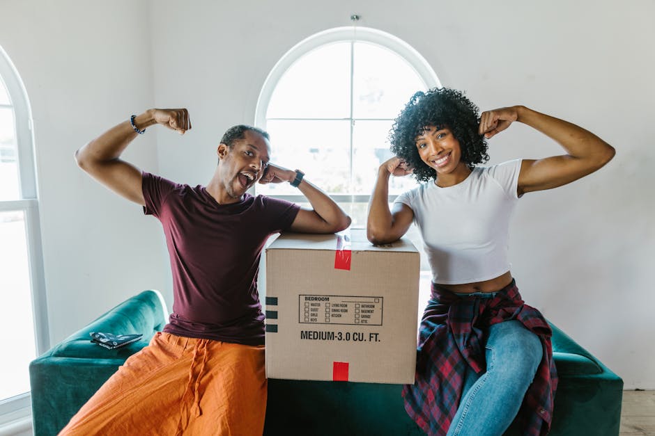 A man and woman indoors, sitting on a teal sofa with a cardboard box between them, during a home relocation process. The man, wearing a maroon t-shirt and orange pants, is smiling widely and flexing his biceps, with his left arm resting on the box. The woman, dressed in a white crop top and blue jeans with a red plaid shirt tied around her waist, is also smiling and flexing her right arm. The cardboard box has printed labels indicating it contains a medium-sized bedroom item with a capacity of 3 cubic feet. Behind them, a large arched window allows natural light into the room, highlighting the clean, modern interior with white walls. Various packing materials, including plastic wrapping, are visible, and a black remote control sits on the sofa. This image captures the packing and moving preparations in a residential setting, illustrating the involvement of professionals such as Man With a Van Holloway in house removals and furniture transport.