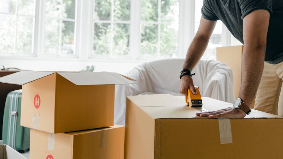 A person measuring a large cardboard box with a tape measure during a home relocation in a well-lit room with large windows showing greenery outside. The individual is wearing a dark t-shirt, beige pants, and a wristwatch, and is leaning over the box with one hand resting on it while the other holds the tape measure to check its dimensions. Several other cardboard boxes, some sealed with packing tape, are stacked nearby on the floor, along with a light green suitcase, indicating packing and moving preparations. A white sofa with a slipcover is visible in the background, along with soft natural daylight illuminating the space, which appears to be part of a residential interior. This scene reflects the loading process typical of house removals, demonstrating the careful packing, measuring for transportation, and organization involved in professional furniture transport and home relocation services, as provided by Man With a Van Holloway.