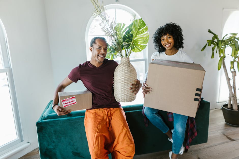 A man and woman indoors, sitting on a teal sofa with a cardboard box between them, during a home relocation process. The man, wearing a maroon t-shirt and orange pants, is smiling widely and flexing his biceps, with his left arm resting on the box. The woman, dressed in a white crop top and blue jeans with a red plaid shirt tied around her waist, is also smiling and flexing her right arm. The cardboard box has printed labels indicating it contains a medium-sized bedroom item with a capacity of 3 cubic feet. Behind them, a large arched window allows natural light into the room, highlighting the clean, modern interior with white walls. Various packing materials, including plastic wrapping, are visible, and a black remote control sits on the sofa. This image captures the packing and moving preparations in a residential setting, illustrating the involvement of professionals such as Man With a Van Holloway in house removals and furniture transport.