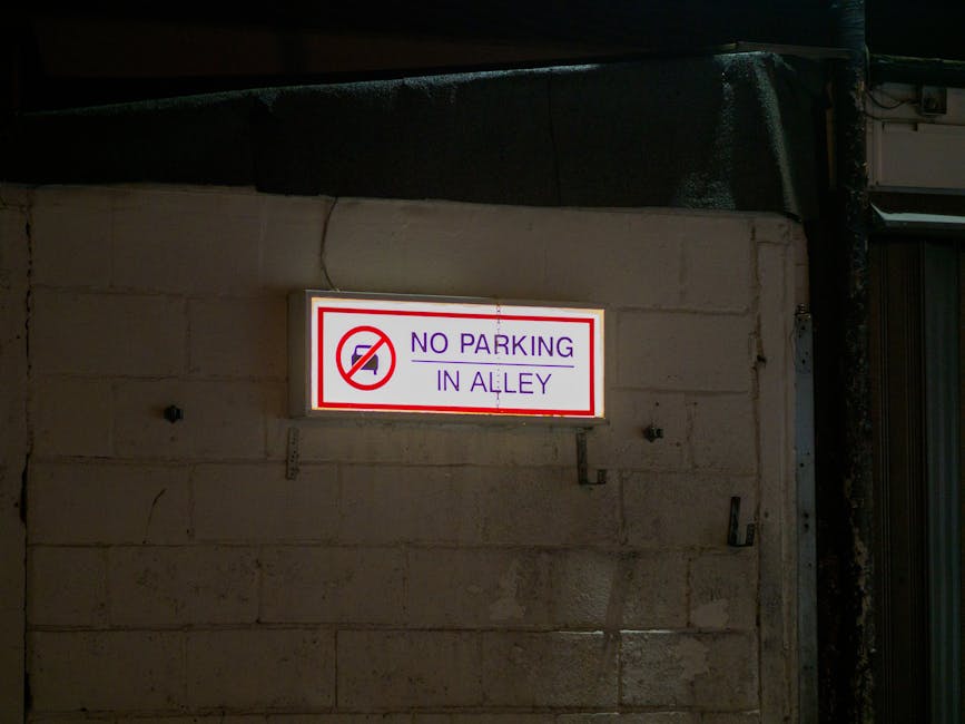 A pair of red Cheltenham Borough Council alcohol-free zone signs attached to a metal pole on a street pavement, with one sign partially overlapping the other. The signs display white text warning that